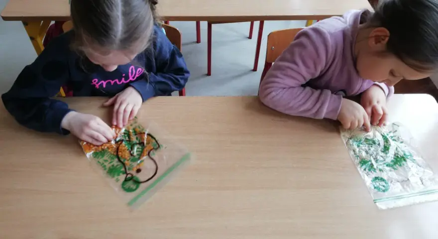 Dwie dziewczynki bawią się materiałami sensorycznymi w woreczkach na drewnianym stole.