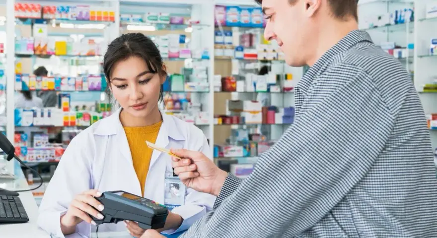 Klient płaci kartą w aptece, gdzie farmaceutka obsługuje terminal. W tle widać półki z lekami, a na nich produkt z napisem ursopol.