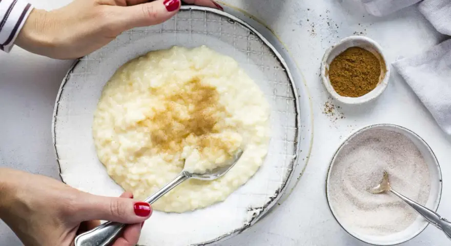 Cremiger Milchreis mit Vanillepudding, bestreut mit Zimt. Eine Hand mit roten Nägeln hält einen Löffel bereit.