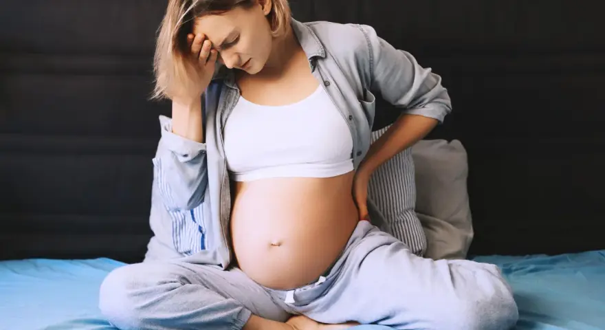 Ciężarna kobieta siedzi na łóżku, trzymając się za głowę i plecy. Odczuwa ból krocza w ciąży, co widać po jej wyrazie twarzy.