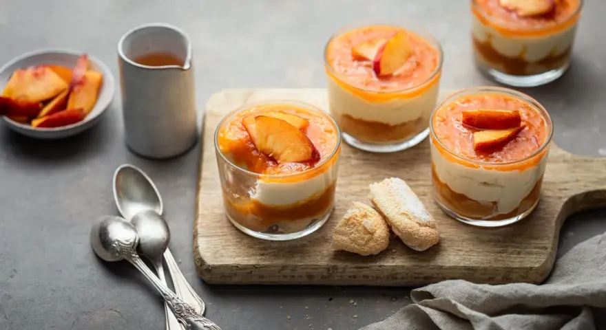 Delizioso tiramisù alle pesche in coppette, con pesche fresche e savoiardi su tagliere di legno.
