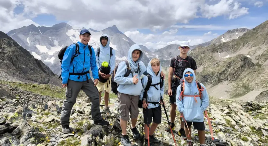 Groupe d'enfants et d'un adulte en randonnée en montagne, souriants, avec des sacs à dos et des bâtons de marche.