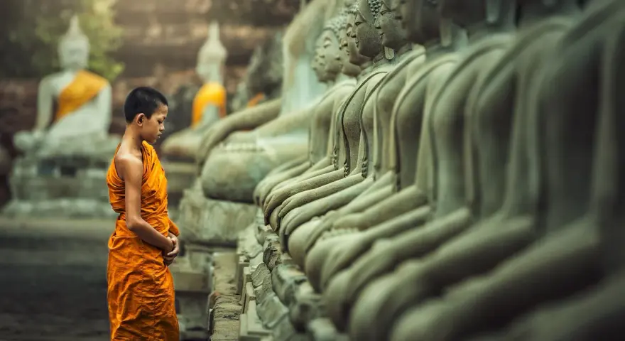 A young monk in orange robes stands before a row of Buddha statues, a scene evoking the ancient roots of what is the oldest religion in the world.