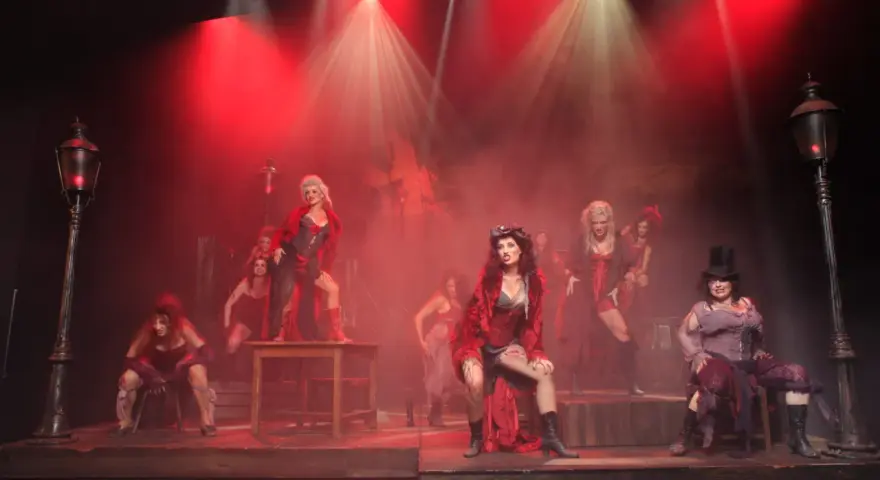 Scena z musicalu "Les Miserables" z kobietami w strojach z epoki, w czerwonym świetle i dymie.