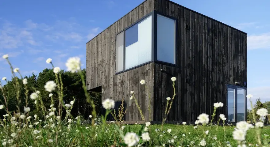 Maison moderne au bardage en bois noir, résultat d'un traitement bois brulé, entourée de fleurs blanches et d'arbres.