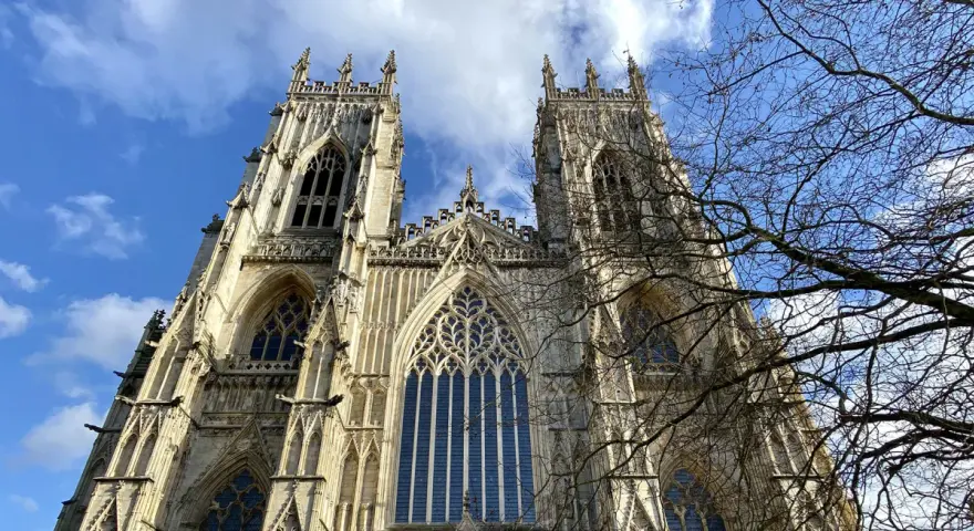 The majestic York Minster, a Gothic cathedral, stands tall against a bright blue sky with scattered clouds. Bare tree branches frame the intricate stonework and towering spires.