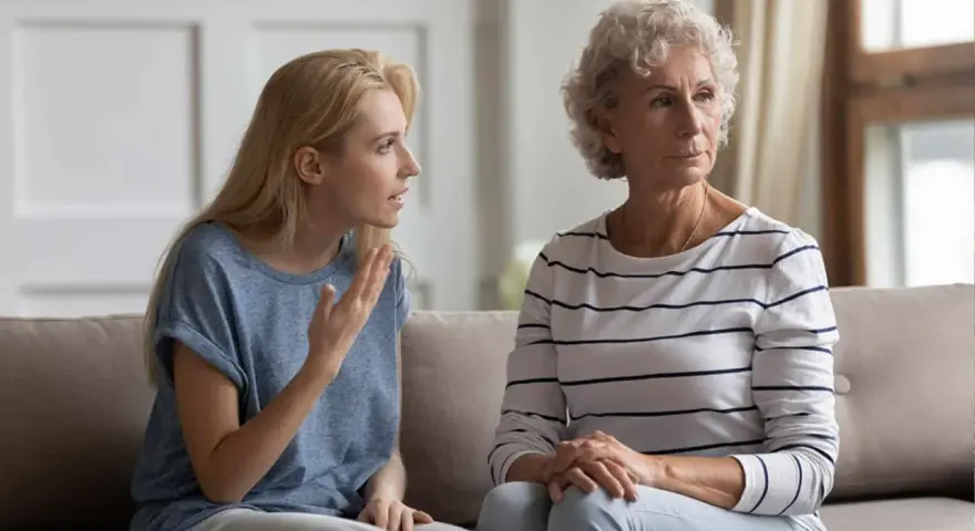 Junge Frau spricht vorwurfsvoll zu ihrer Mutter, die abgewandt sitzt. Die Mutter beleidigt die erwachsene Tochter.