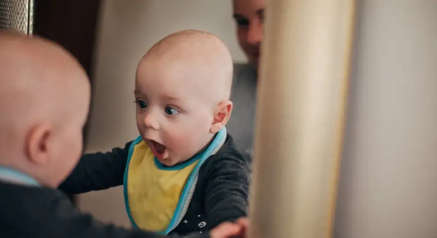 Ein Baby mit gelbem Lätzchen schaut überrascht in den Spiegel. Es ist fasziniert von seinem Spiegelbild, ein Moment, wann sagen Kinder "Ich".