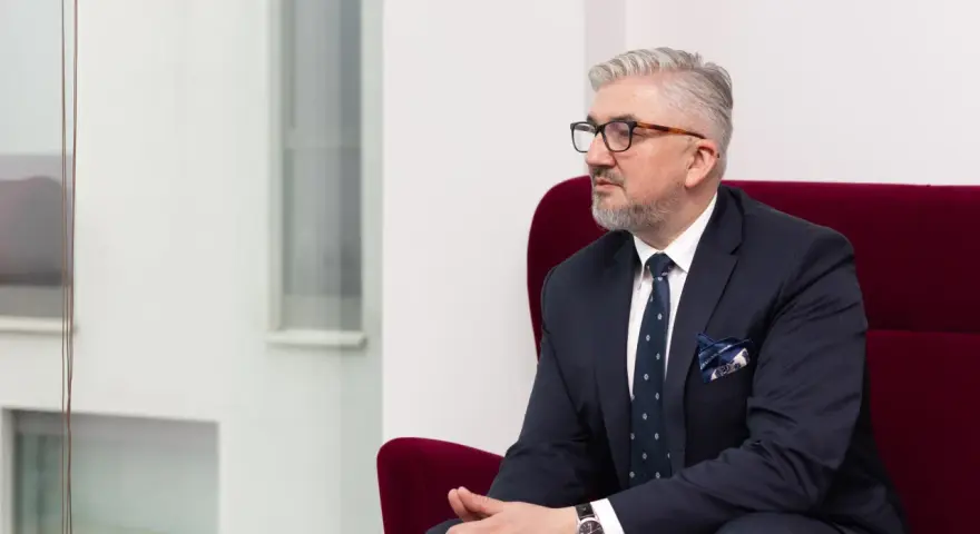 Mężczyzna w garniturze i okularach siedzi na czerwonym fotelu, patrząc w bok.