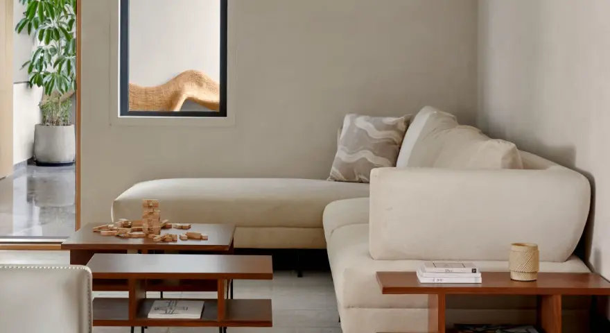 Sofá modular beige, mesa de centro de madera con juego de Jenga, planta en maceta y ventana con detalle decorativo. Ambiente de estilo cozy.