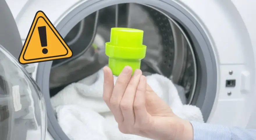 Main dans la machine à laver, tenant un bouchon vert pour le bac adoucissant. Un panneau d'avertissement jaune est visible.