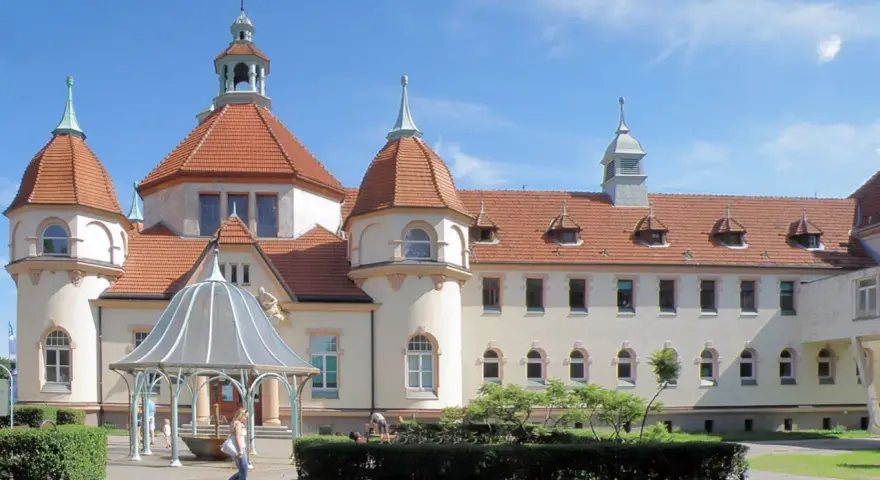 Elegancki zakład balneologiczny Sopot z charakterystycznymi wieżyczkami i czerwonym dachem, otoczony zielenią.