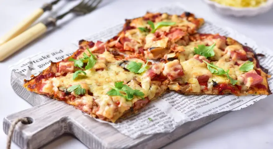 Pyszna pizza na spodzie z kalafiora keto, z szynką i serem, udekorowana świeżą pietruszką.