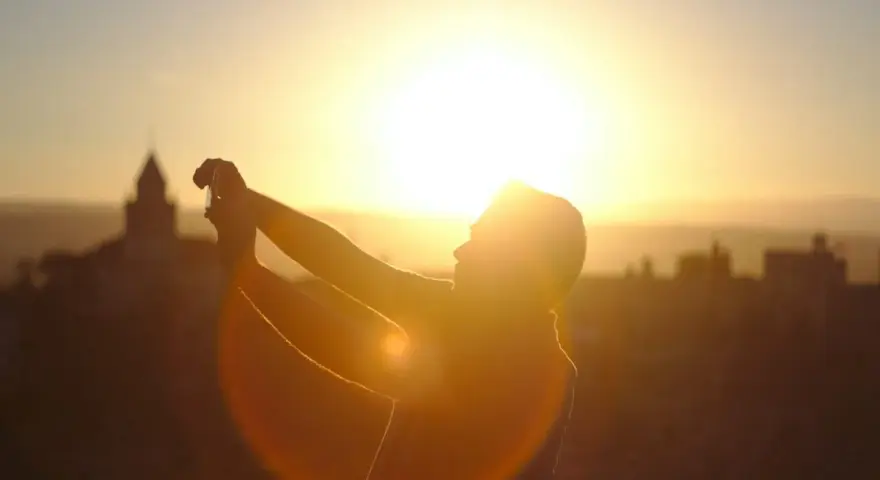 Silhouette d'une personne prenant une photo contre jour avec son téléphone, le soleil éclatant à l'horizon derrière des bâtiments.