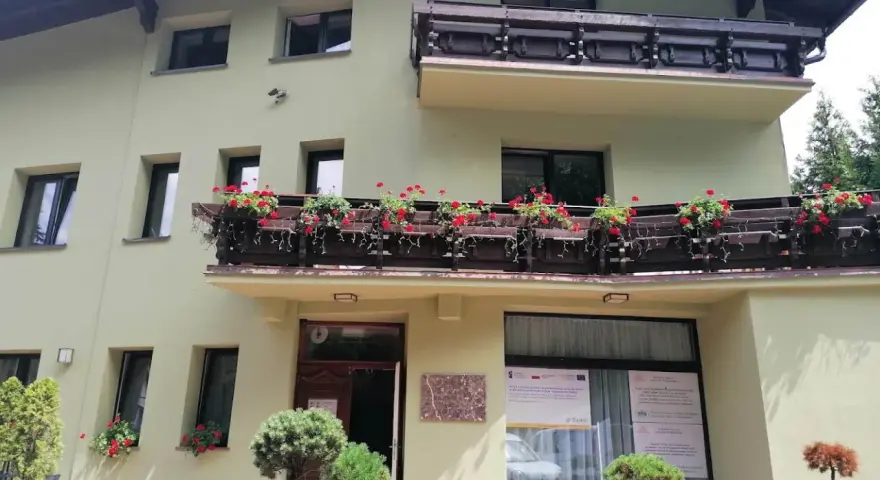Jasnozielony hotel na błoniach z balkonem pełnym czerwonych kwiatów.