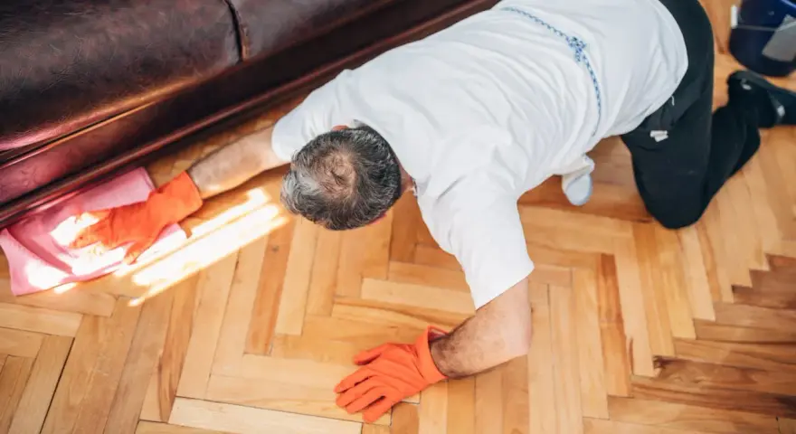Man in orange gloves cleaning wooden floor with a pink cloth.