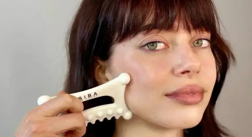 A woman with bangs and green eyes performs a facial fascia massage using a white gua sha tool.