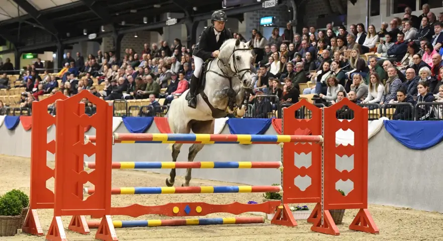 Un cheval blanc, un véritable con air etalon, saute une barrière colorée sous les regards d'une foule attentive.