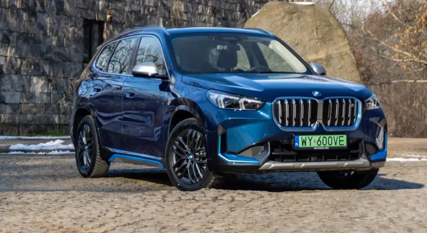 Niebieskie BMW iX1 stoi na kamienistej nawierzchni przed kamienną ścianą.