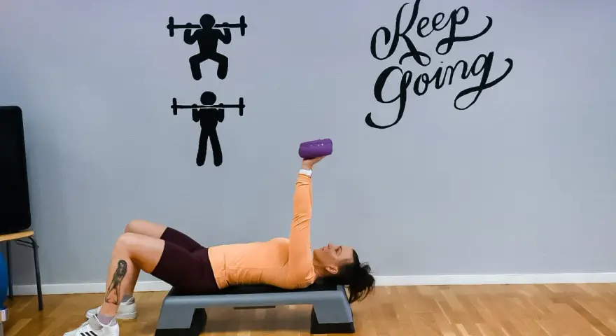 Kvinna tränar med en lila hantel på en stepbräda. Väggen har texten "Keep Going" och silhuetter av styrkelyft.