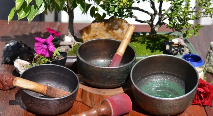 Tres cuencos tibetanos para limpiar piedras, con mazos de madera y detalles ornamentales, junto a un bonsái y flores.