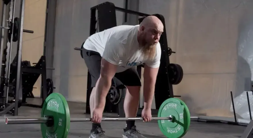 Man med skägg utför raka marklyft med hantlar i ett gym.