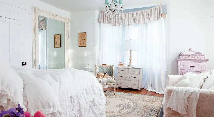 Dormitorio con cama blanca y volantes, espejo grande, cómoda antigua y sillón. Estilo shabby chic.
