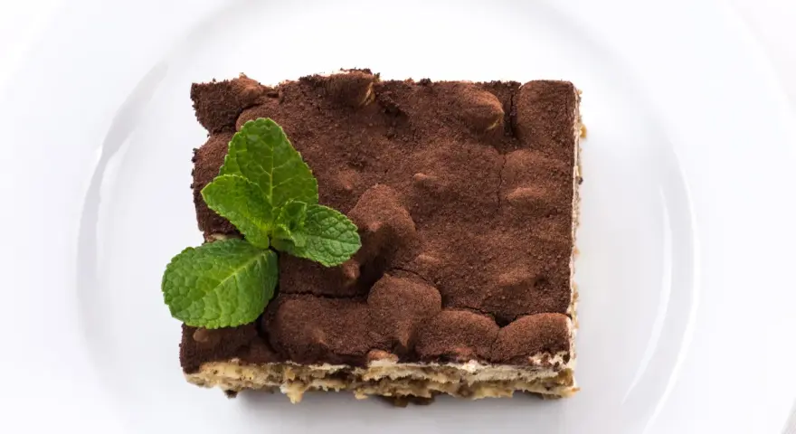 Pyszne tiramisu bez alkoholu, posypane kakao i udekorowane listkami mięty, na białym talerzu.