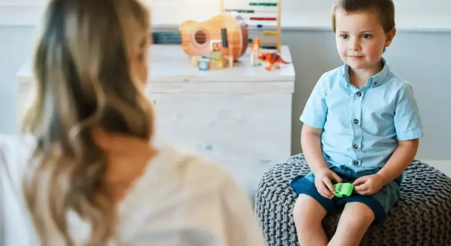 Chłopiec siedzi na pufie naprzeciwko psychoterapeutki, w tle zabawki.