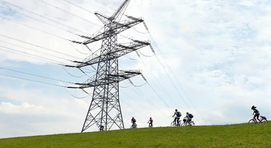 Grupa rowerzystów przemierza zielone wzgórze pod potężną konstrukcją linii 110 kV.