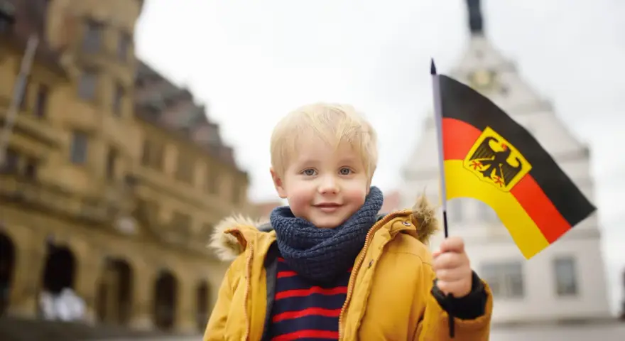 Uśmiechnięty chłopiec z blond włosami trzyma flagę Niemiec. W tle widać zabytkowe budynki, przypominające te, które można spotkać w niemieckich miasteczkach, gdzie znajdują się szkoły.