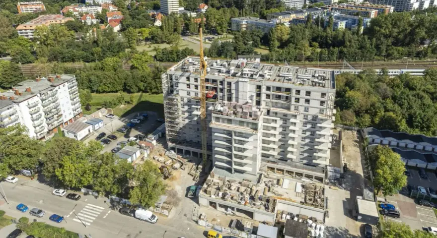 Budowa bloku mieszkalnego w Kołobrzegu. Widok z lotu ptaka na plac budowy, dźwig, samochody i okoliczne budynki. Ceny mieszkań Kołobrzeg mogą być różne.