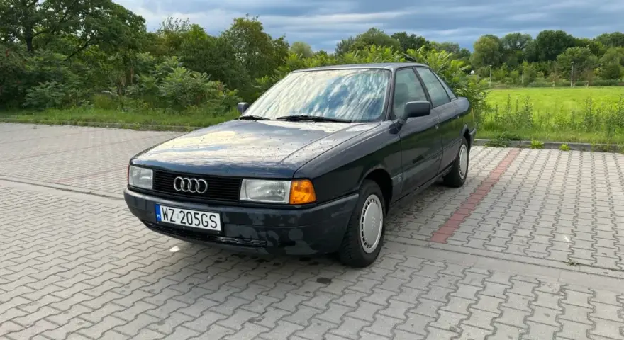 Ciemny Audi 80, klasyk z lat 90., idealny dla szukających aut do 5 tys. zł. Zaparkowany na kostce brukowej, otoczony zielenią.