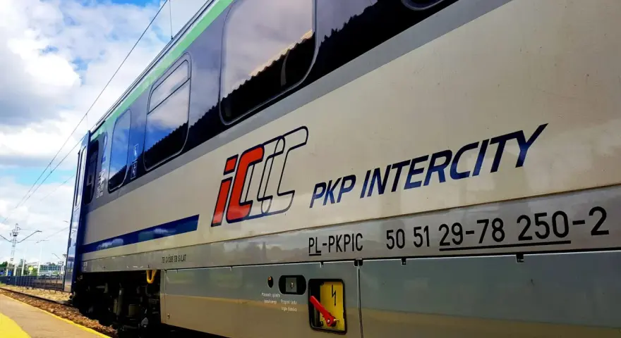Pociąg PKP Intercity z napisem "Warszawa Poznań" na tle błękitnego nieba z chmurami.