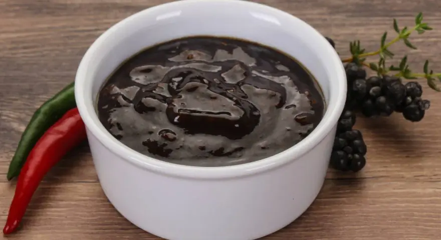 Gęsty, ciemny sos pieprzowy w białej miseczce, obok czerwona i zielona papryczka chili oraz gałązka jeżyn.