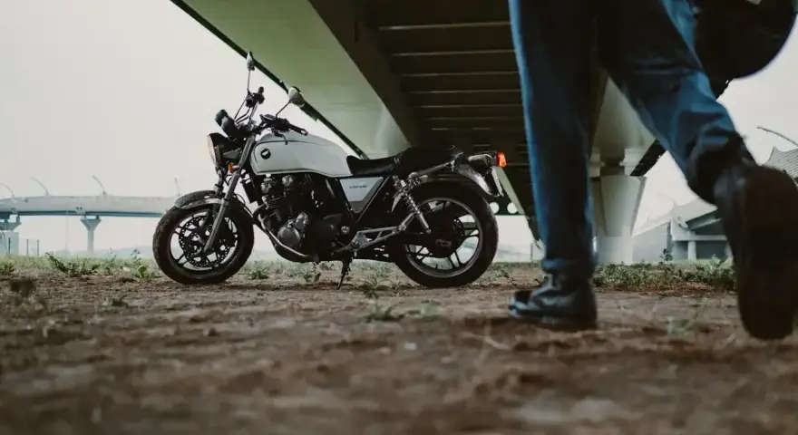 Biały motocykl klasyczny, kategoria A2, pod mostem. Obok przechodzi motocyklista z kaskiem.