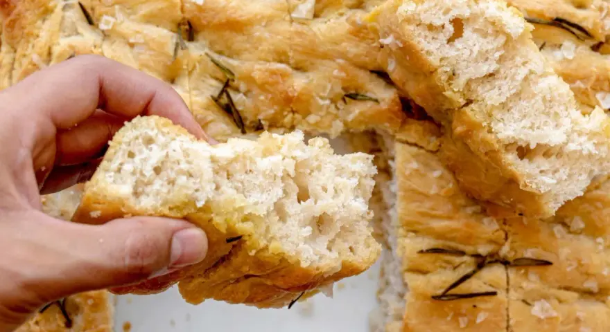 Mano rompiendo un trozo de focaccia casera con romero y sal gruesa. ¡Una delicia de esta focaccia receta!