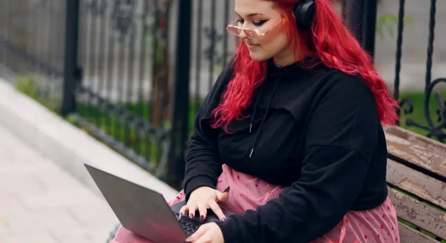 Młoda babista z ognistymi włosami i słuchawkami na uszach pracuje na laptopie, siedząc na ławce w parku.