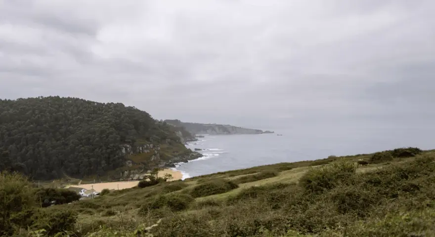 Paisaje costero con acantilados verdes y mar gris. Ideal para senderismo en Villaviciosa, con una playa dorada y una casa blanca.