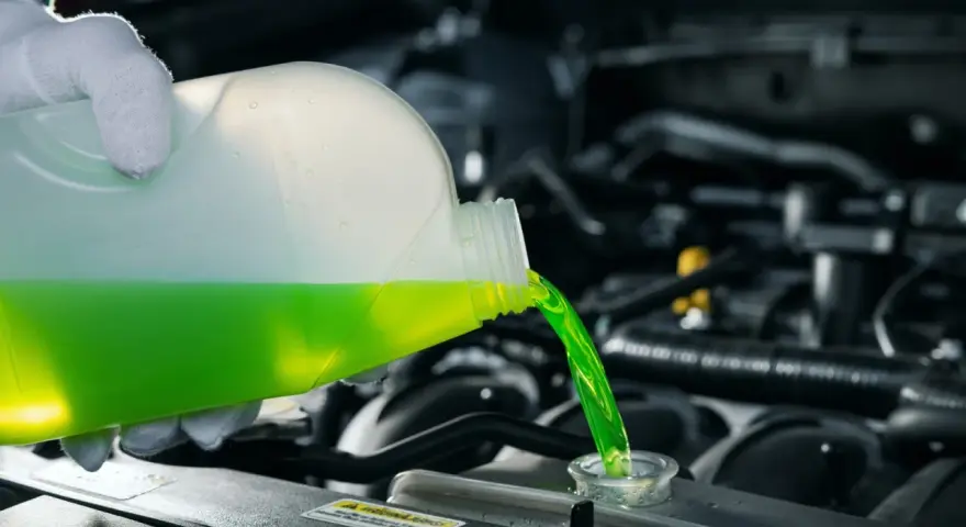 A gloved hand pours green antifreeze and coolant into a car's engine.