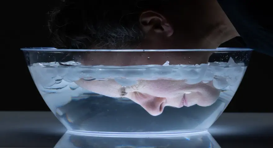A person submerges their face in a clear bowl filled with water and ice cubes for a facial ice bath.