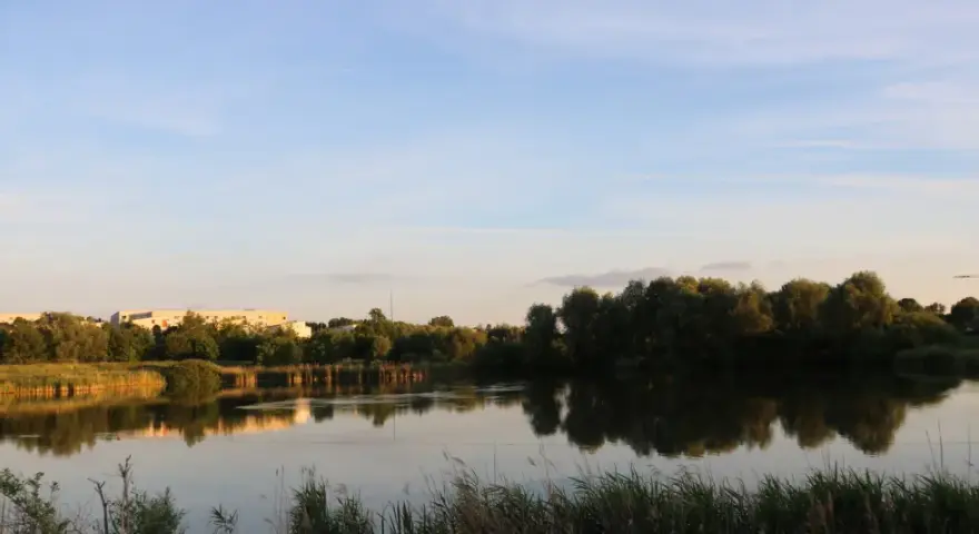 Spiegelglatte Wasserfläche am Wuhletal Wanderweg, reflektiert Bäume und Gebäude. Ein rotes Haus sticht hervor.