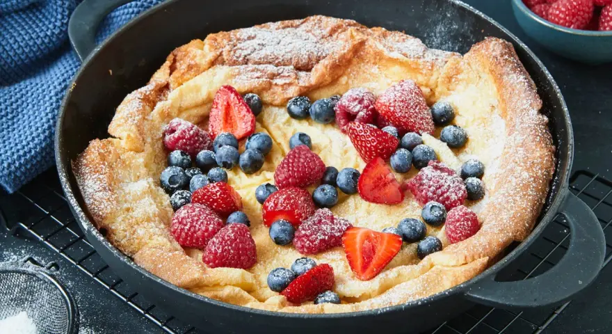 Ein goldbrauner Dutch Baby mit Puderzucker und frischen Beeren, serviert in einer gusseisernen Pfanne.