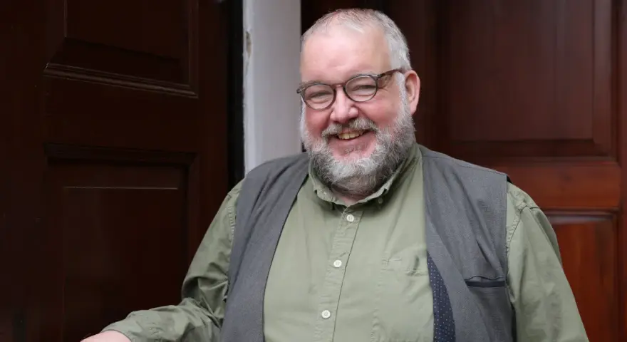Ein freundlicher evangelischer Pfarrer mit Brille und Bart lächelt in die Kamera, vor einer dunklen Holztür stehend.