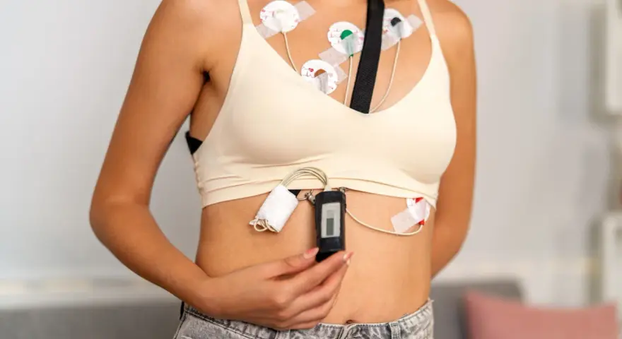 Kobieta z przyklejonymi elektrodami do klatki piersiowej, podłączonymi do rejestratora. To holter ekg, monitorujący pracę serca.