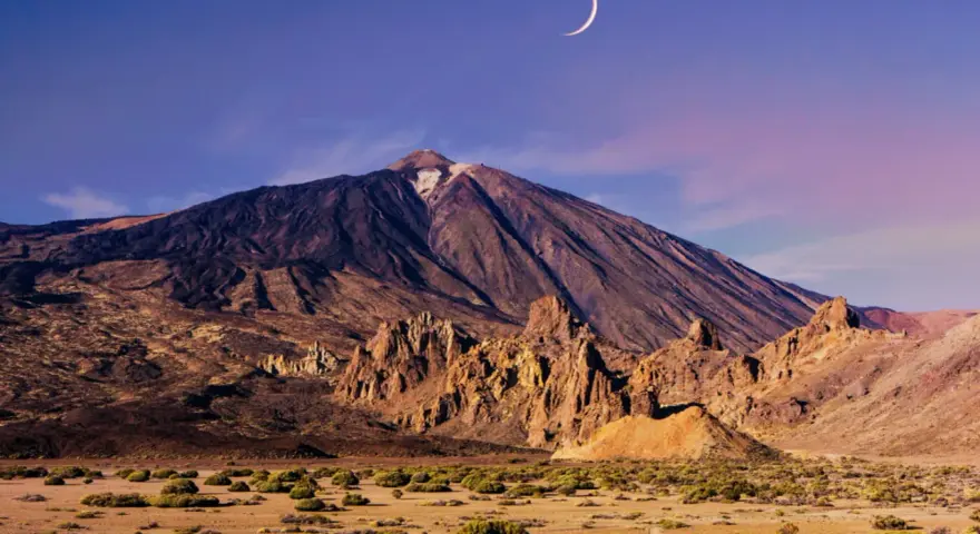 Krajobraz w parku narodowym Teide z księżycem w kształcie sierpa na fioletowym niebie. Wulkan góruje nad skalistym terenem.