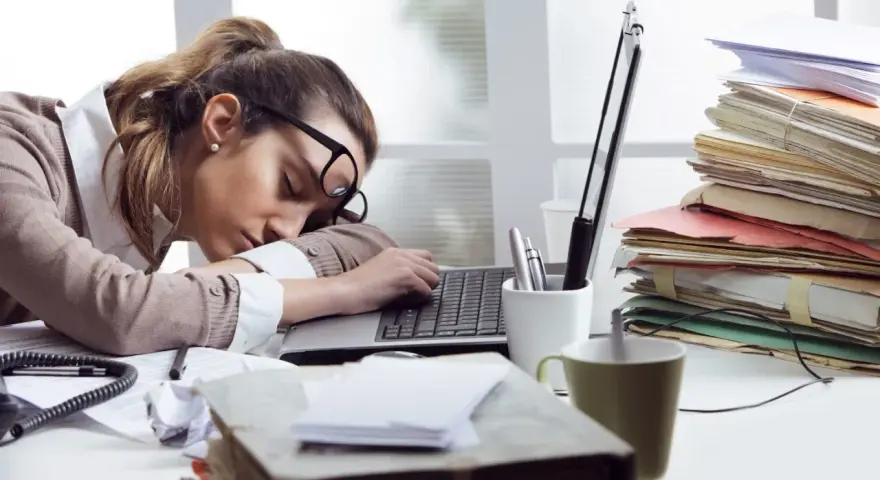 Zmęczona kobieta śpi na laptopie, otoczona stertami dokumentów. To obrazek pokazujący skutki związku z pracoholikiem.