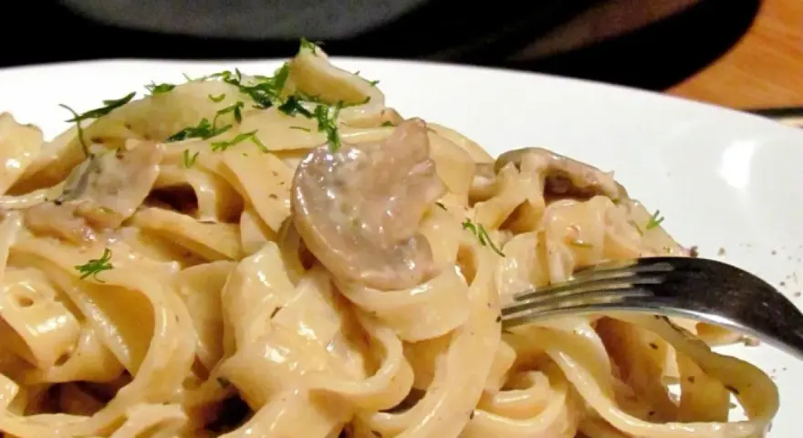 Tagiatelle z grzybami w kremowym sosie, posypane świeżym koperkiem. Widok z bliska na talerz pełen pysznego makaronu.