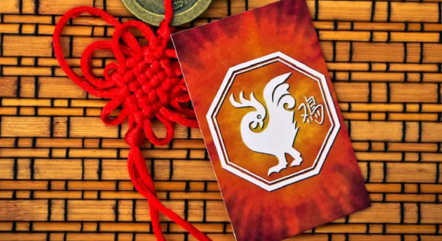 Czerwony chiński węzeł i karta z białym kogutem, symbolem 1993 chińskiego znaku zodiaku, na tle bambusowym.