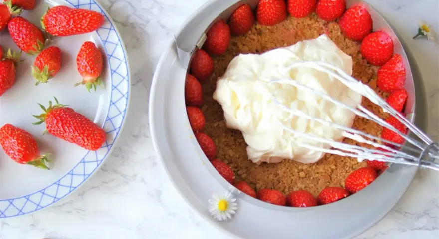 Préparation d'un cheesecake fraise : crème fouettée sur une base de biscuits et fraises fraîches.